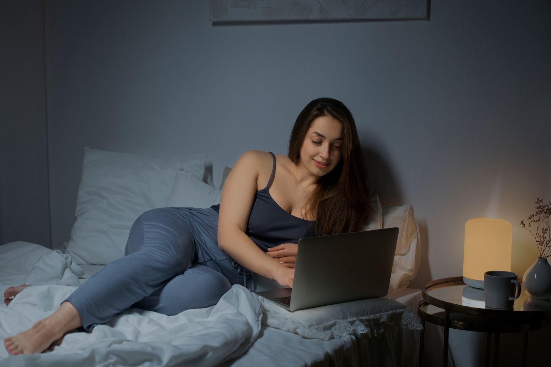 Une femme allongée sur son lit travaille sur un ordinateur portable, avec une lampe de chevet allumée et une tasse sur la table.